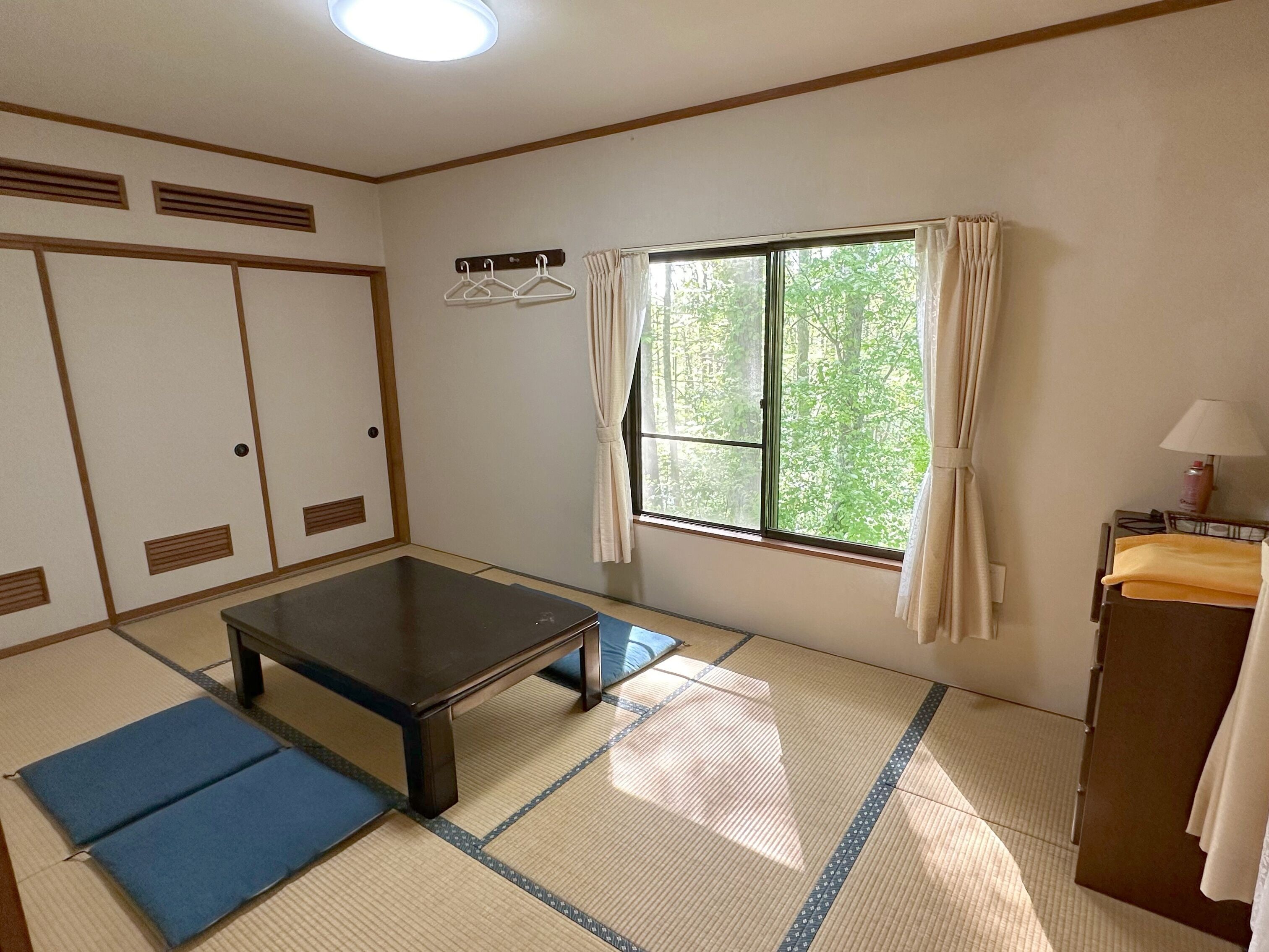 A room with tatami mats, a low table, a window with curtains, a wardrobe, and a small desk with a lamp.