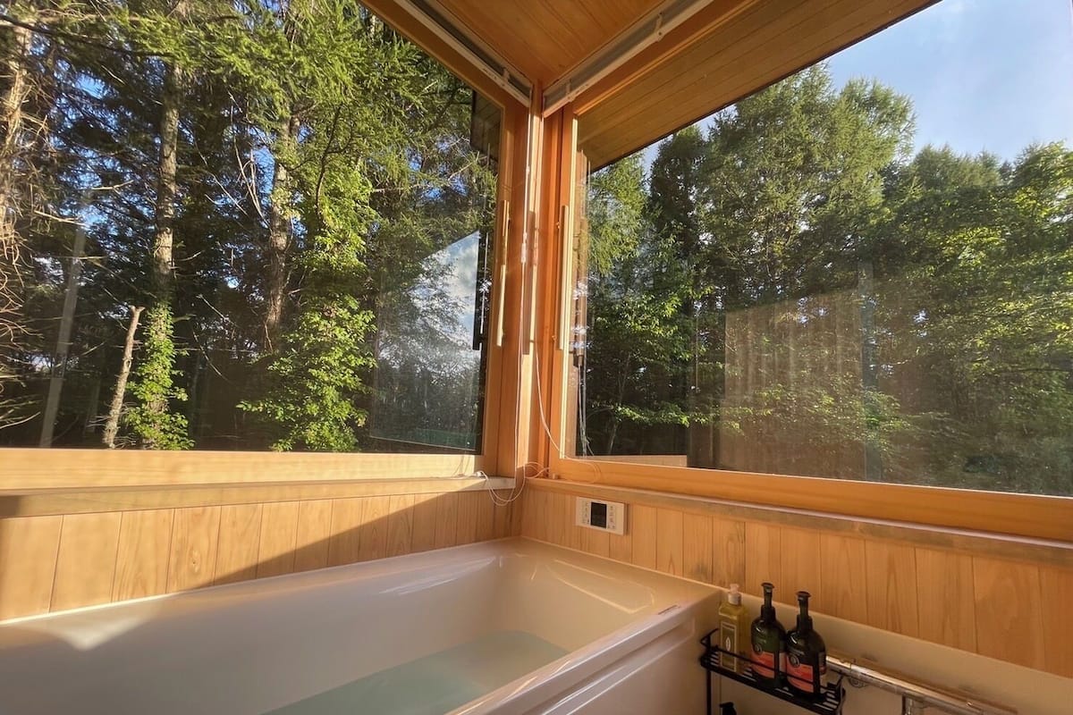A wooden cabin with a large window, a bathtub, and a shelf with bottles.