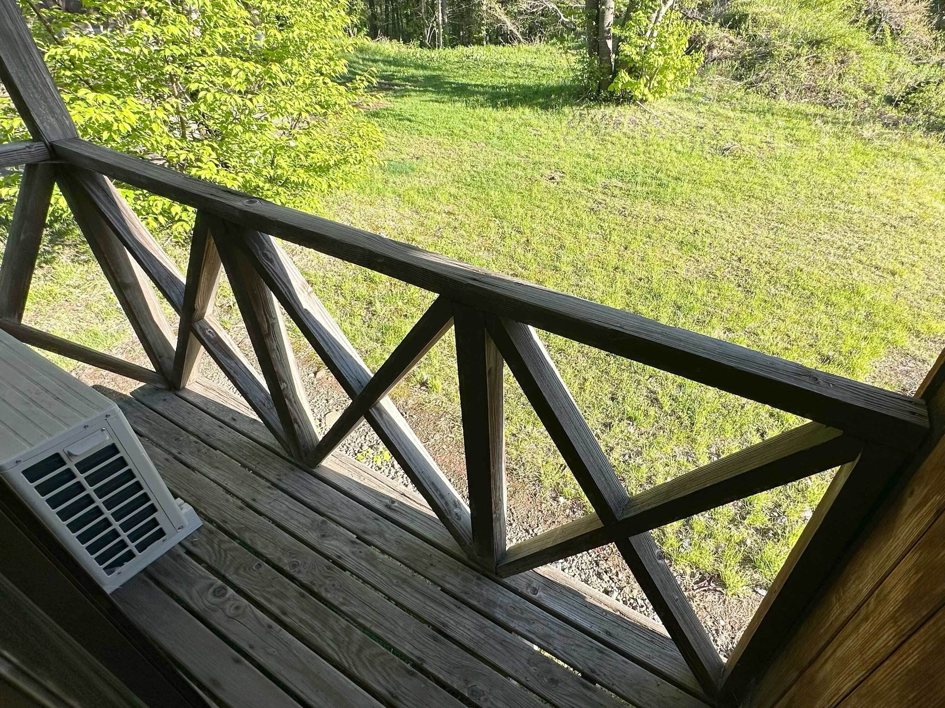 Eine Holzterrasse mit einem Metalllüftungsgitter, mit Blick auf eine Wiese mit Bäumen.