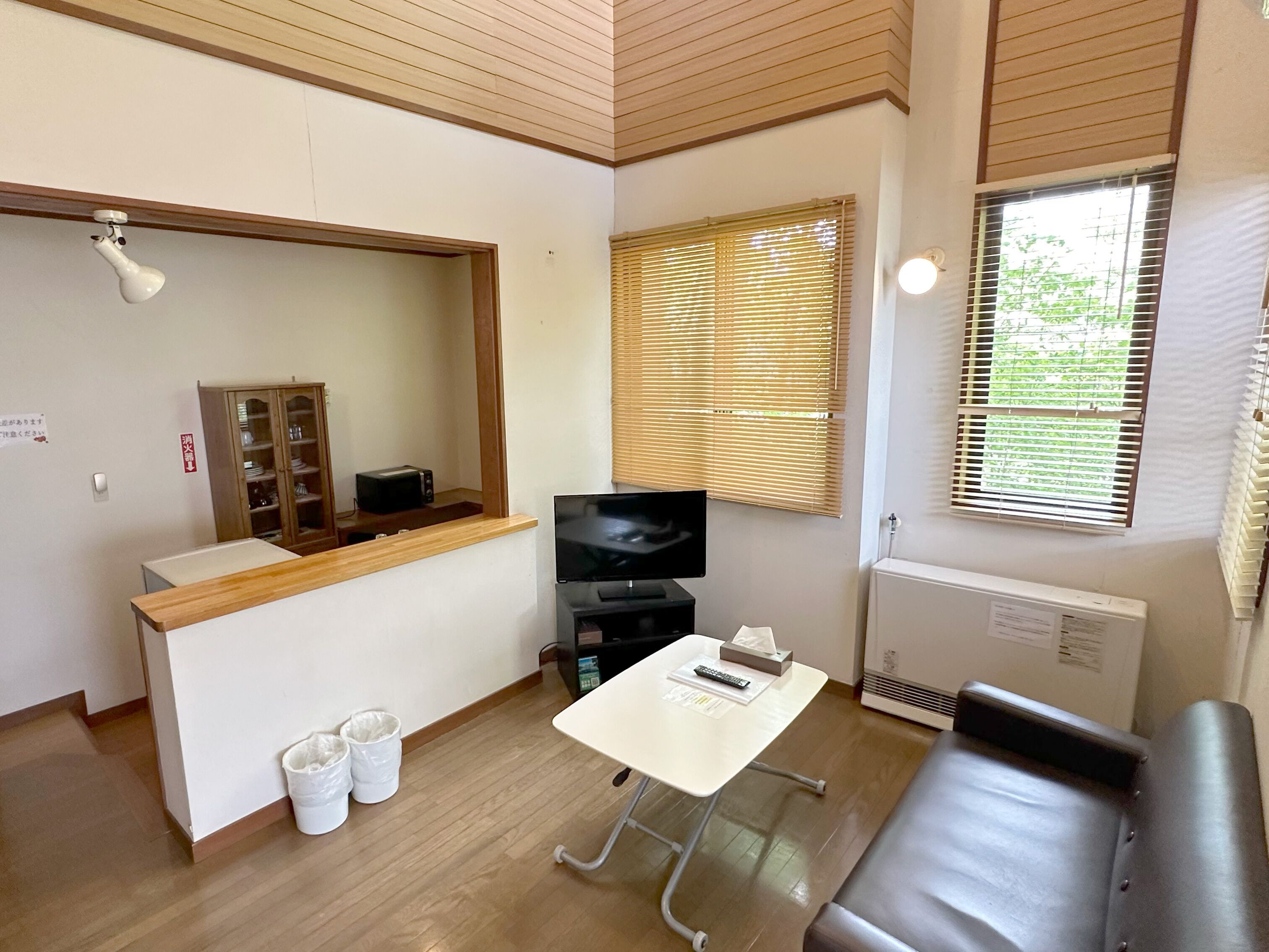 A compact living space with a wooden ceiling, a TV, a small table with a remote, a cushioned bench, and a window with blinds.