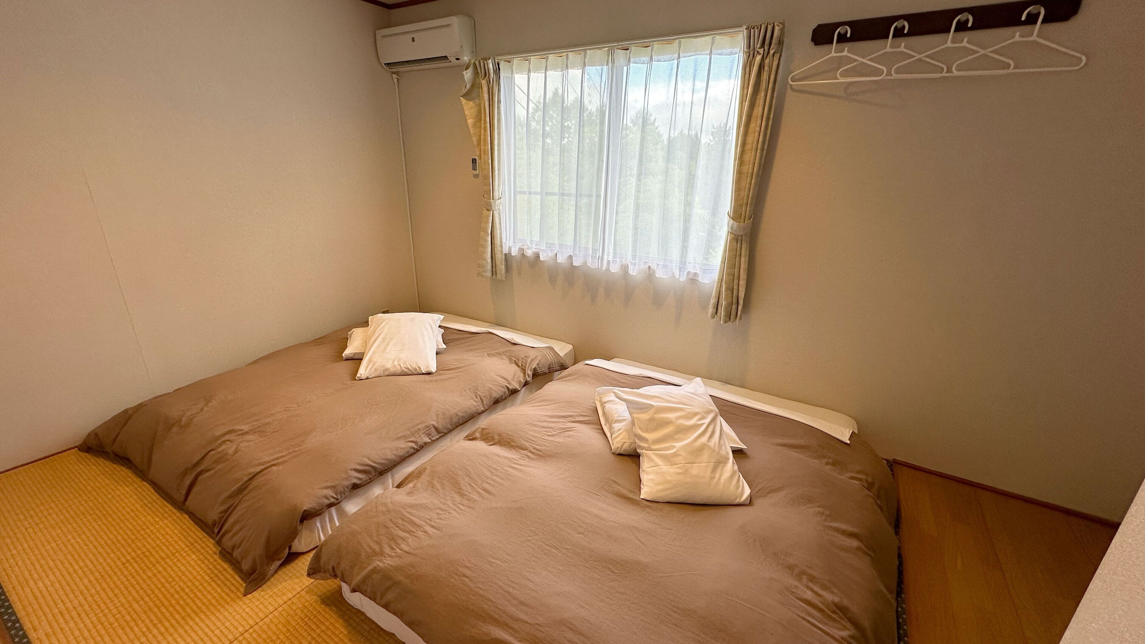 A small, single-room accommodation with two beds, a window with curtains, a wall-mounted air conditioner, and a wooden floor.