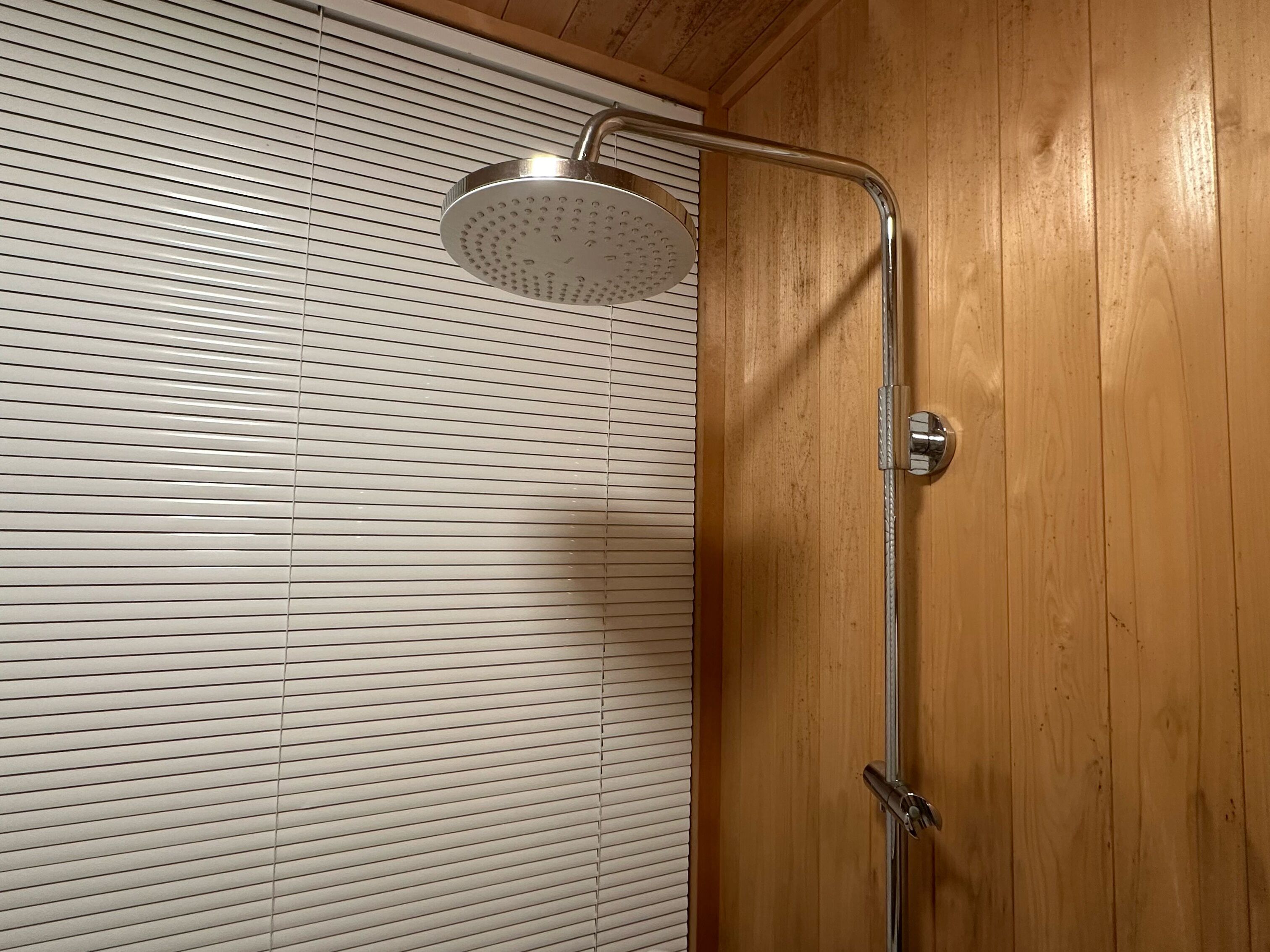 A shower with a curved head and a chrome handle, mounted on a wooden wall with horizontal blinds.