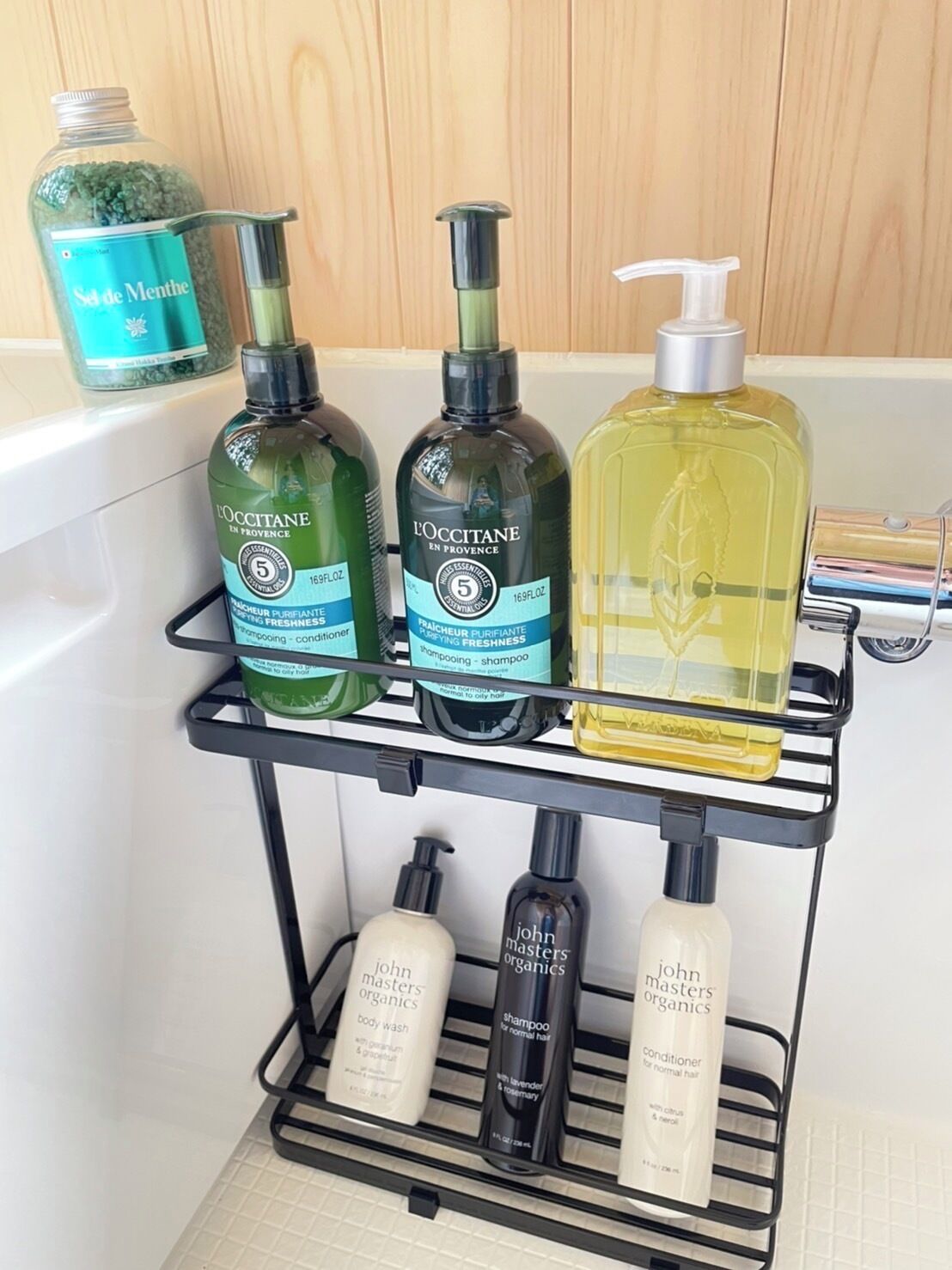 Un estante de baño con varios artículos de aseo personal, incluyendo productos L'OCCITANE, John Masters Organics, y una botella de champú de lavanda y romero.