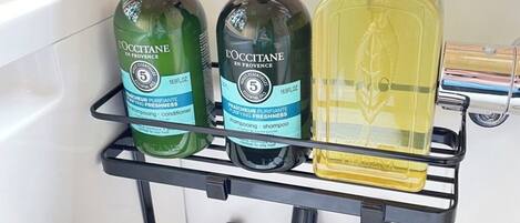 Un estante de baño con varios artículos de aseo personal, incluyendo productos L'OCCITANE, John Masters Organics, y una botella de champú de lavanda y romero.