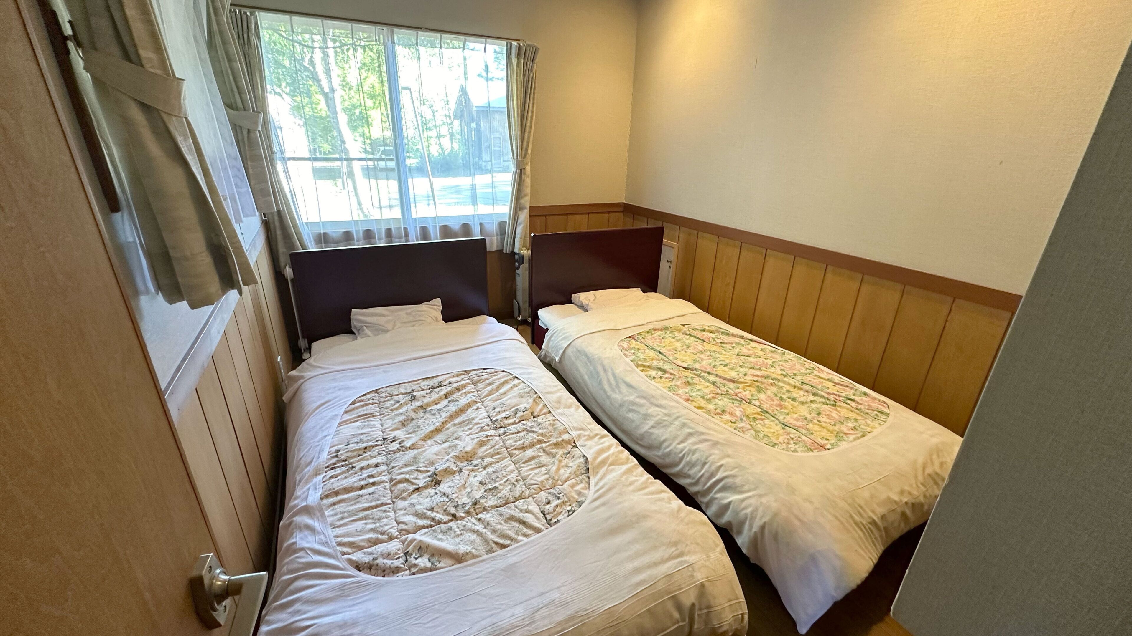 A small room with two single beds, a window with curtains, and a wooden headboard.