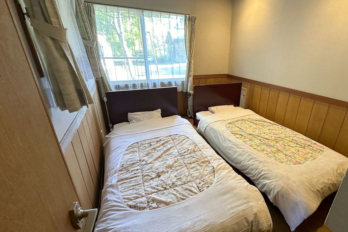 A small room with two single beds, a window with curtains, and a wooden headboard.