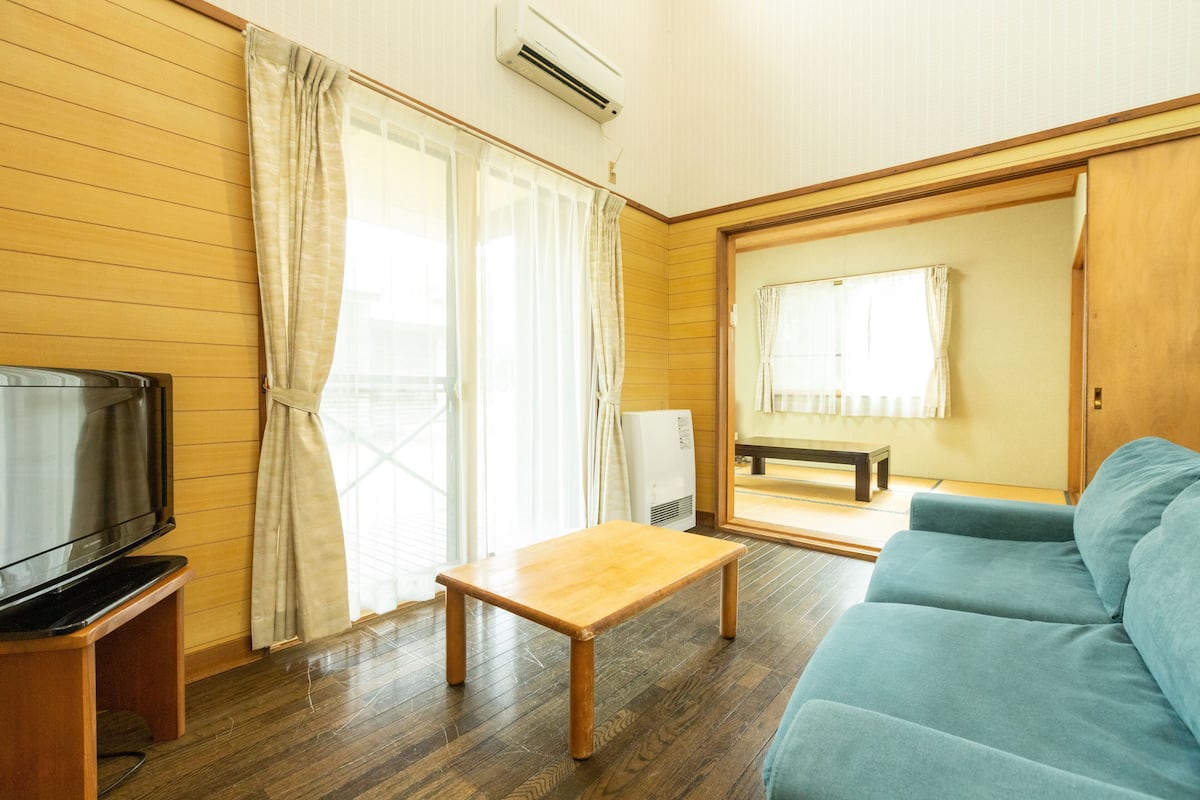 A room with a wooden floor, a blue sofa, a wooden table, a television, and a sliding door leading to another room.