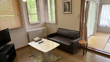 Une pièce comprenant un canapé, une petite table, un téléviseur et une fenêtre avec des stores.
