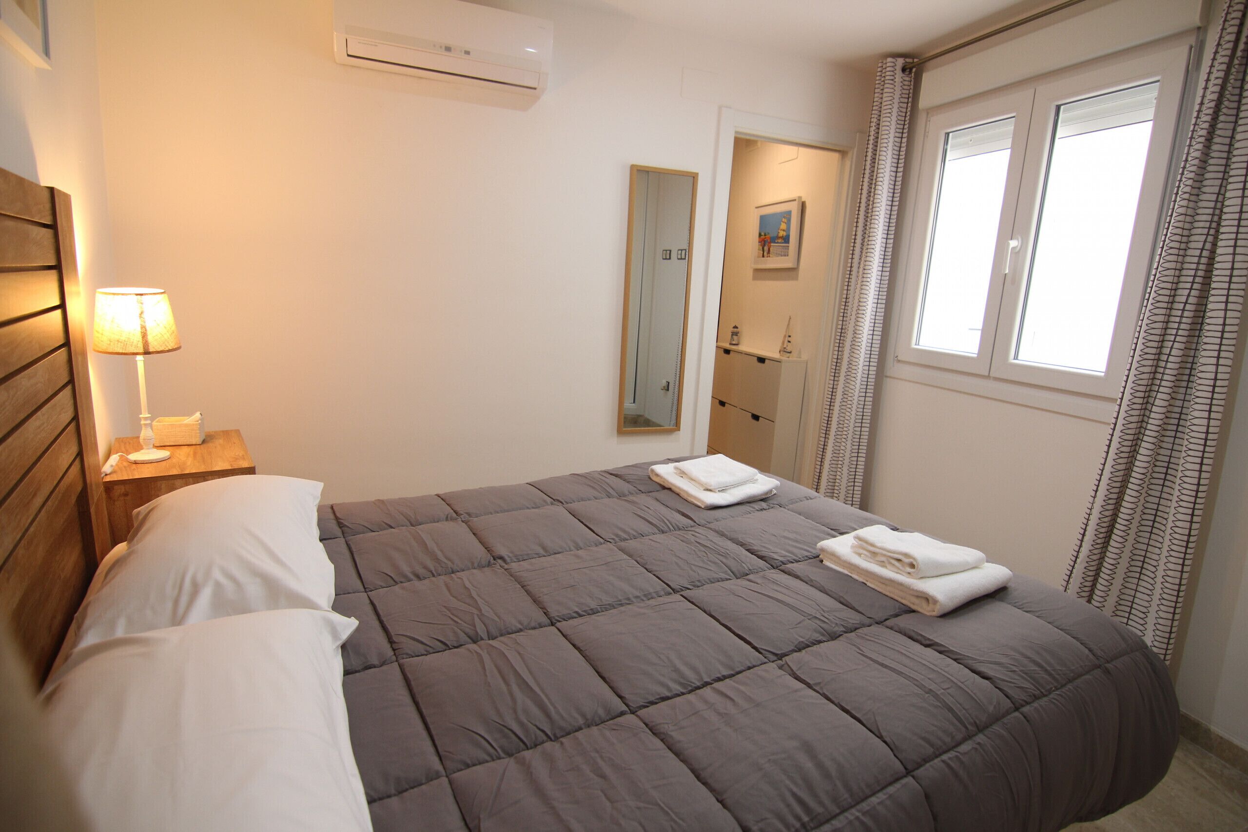 A neatly made bed with a grey duvet, a bedside lamp, and a mirror on the wall.
