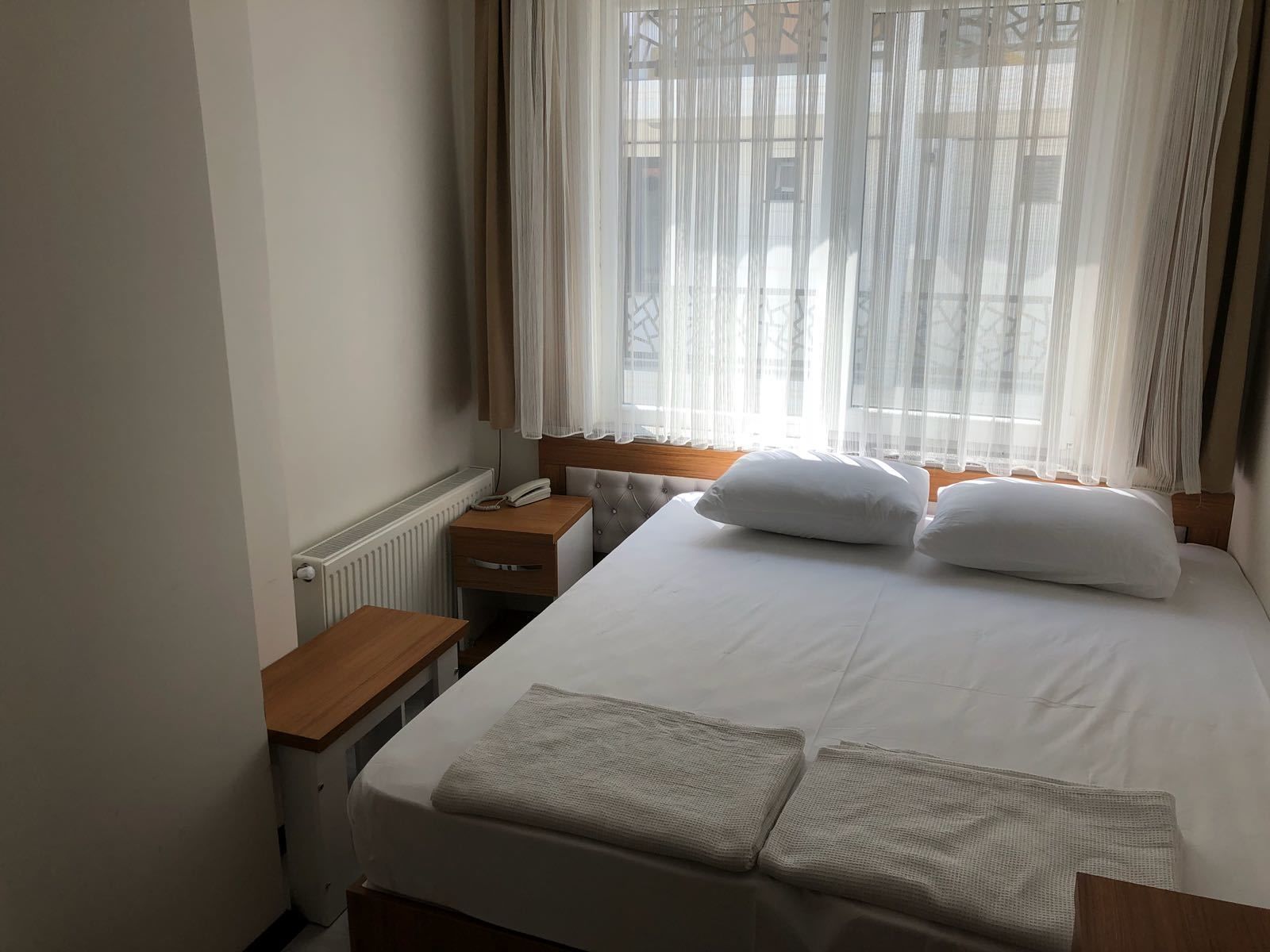 Una habitación de hotel con cama, mesita de noche, lámpara y ventana con cortinas de gasa.