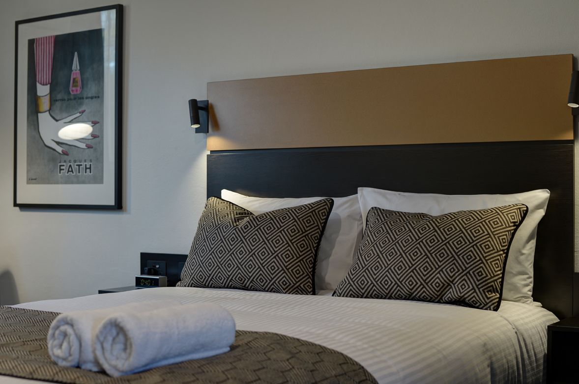 A neatly made bed with patterned pillows, a framed poster on the wall, and a bedside table with a lamp.