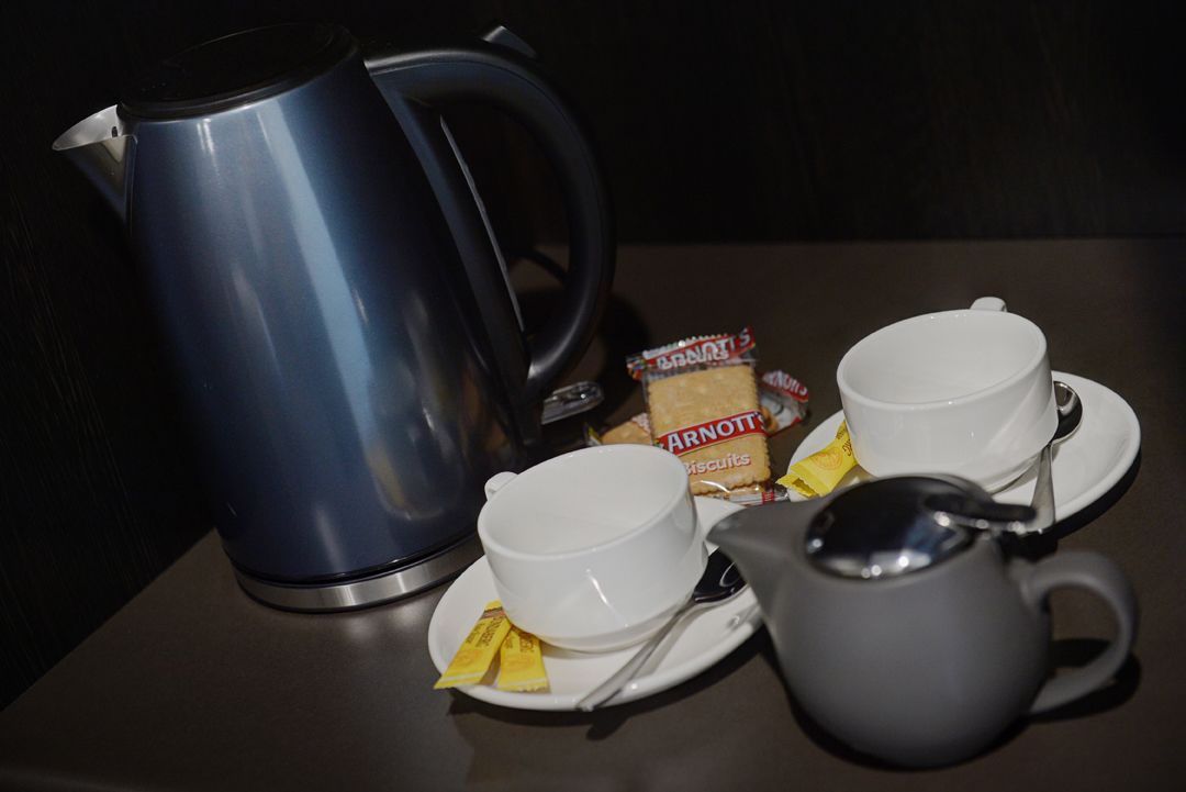 Een tafel met een blauwe elektrische waterkoker, twee witte bekers met schotels, een grijze mok, een lepel en een pakje Arnott's koekjes.