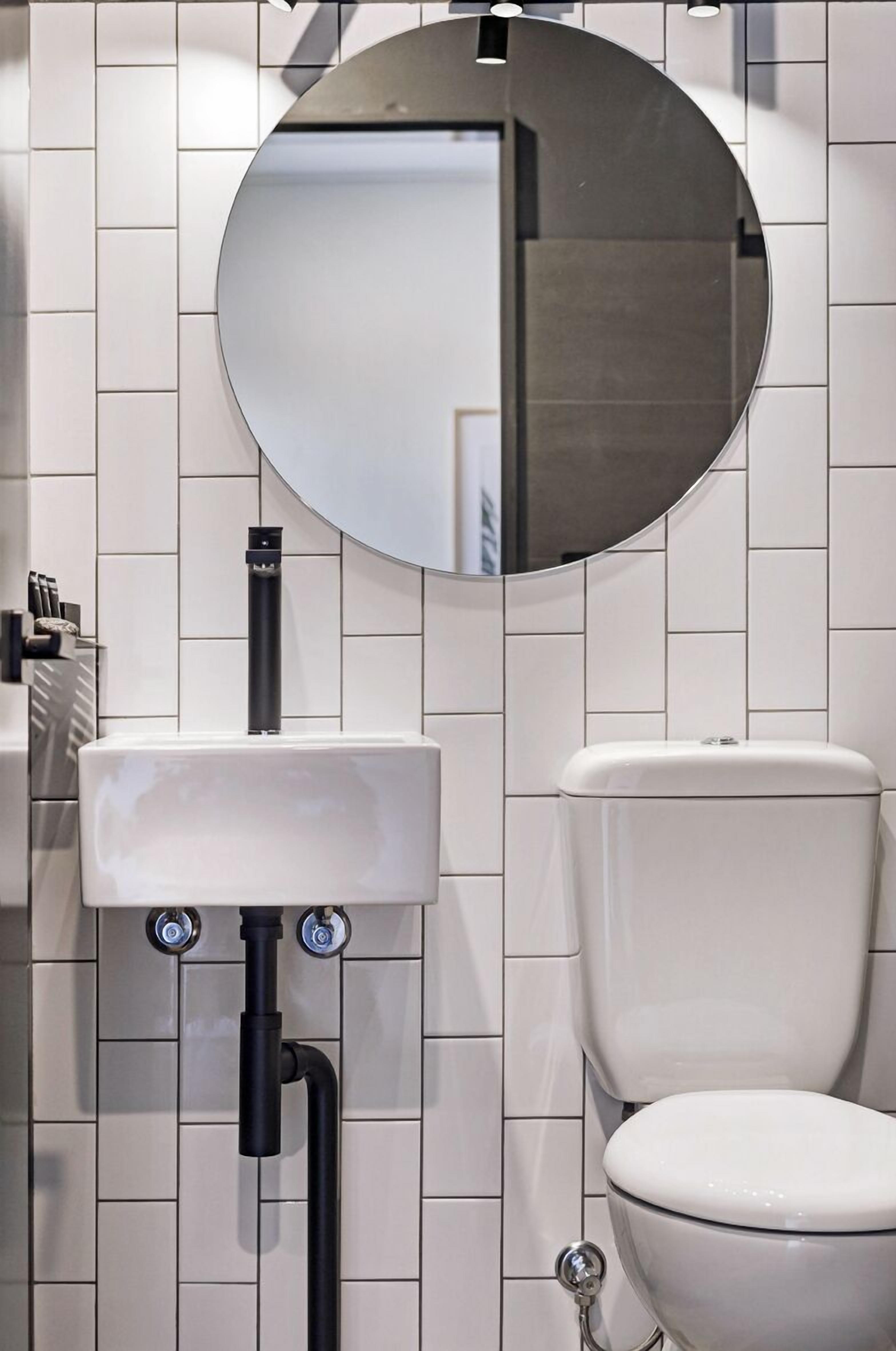 Un baño moderno con pared de azulejos blancos, un espejo redondo, un lavabo blanco y un inodoro.