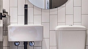 A modern bathroom with a white tiled wall, a round mirror, a white sink, and a toilet.