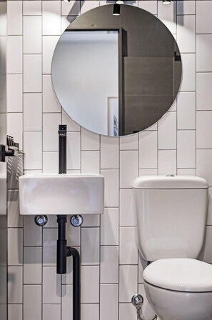 A modern bathroom with a white tiled wall, a round mirror, a white sink, and a toilet.