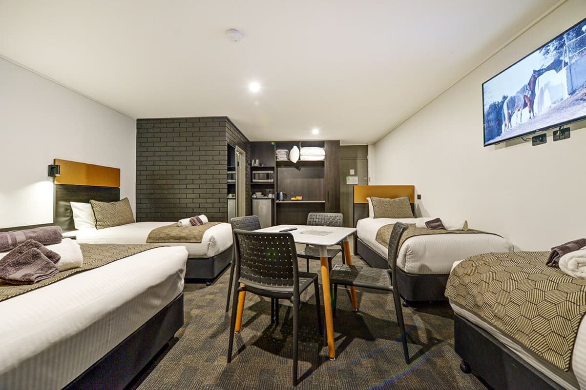A hotel room with two beds, a dining table, a kitchen area, and a TV mounted on the wall.