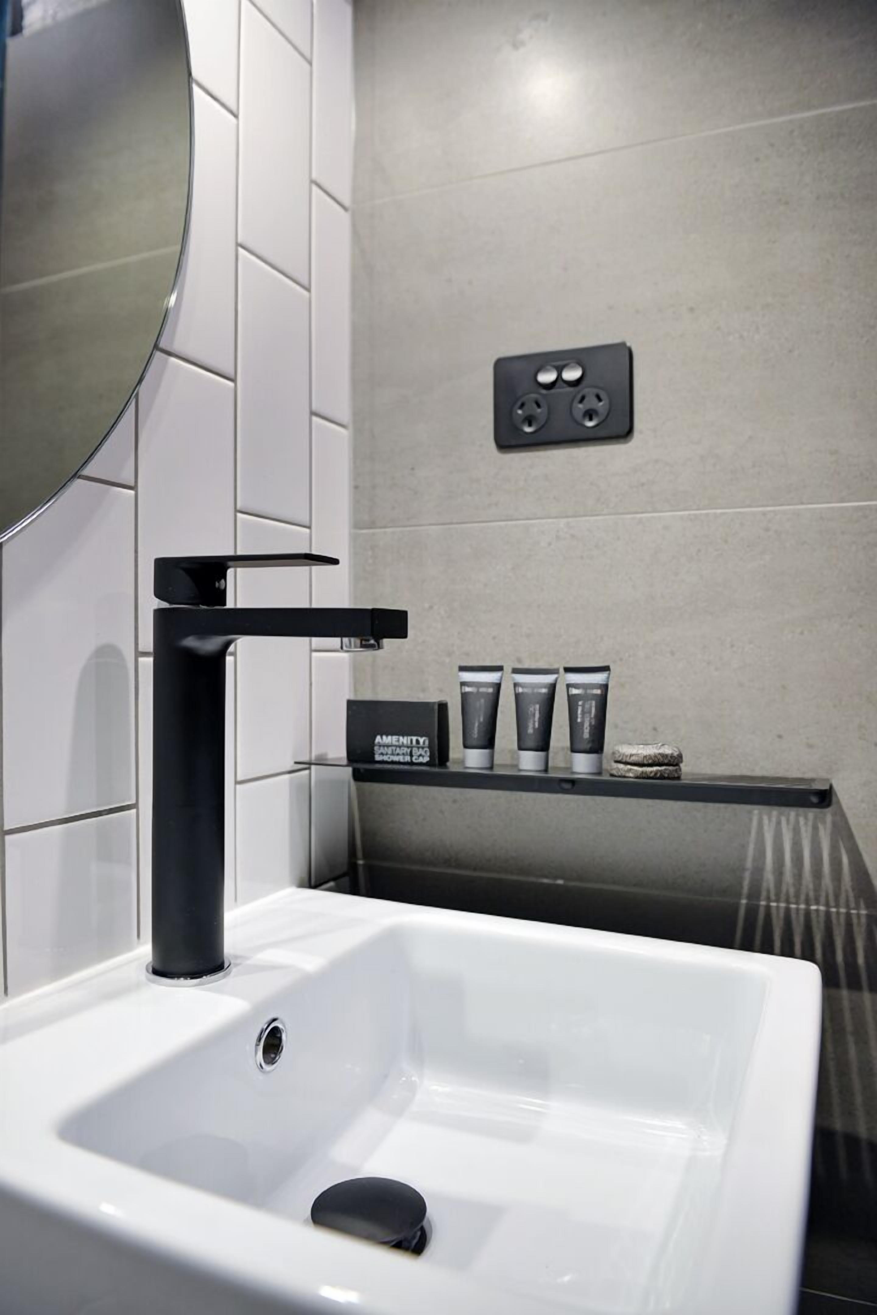 Un baño moderno con un lavamanos blanco, un grifo negro y un espejo. Hay tres artículos de tocador en un estante y un enchufe eléctrico negro en la pared.