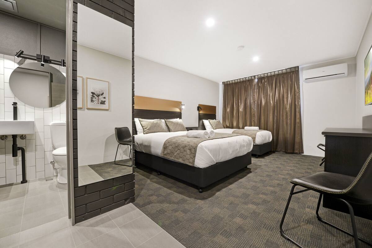 A hotel room with a large bed, a desk, a chair, a mirror, a toilet, and a shower area.