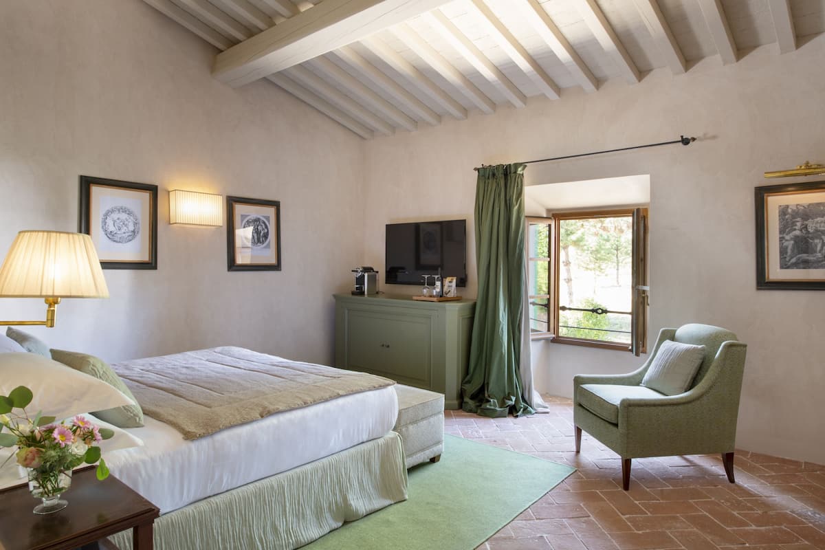 Een slaapkamer met een bed, een stoel, een dressoir, een televisie en een raam met gordijnen.