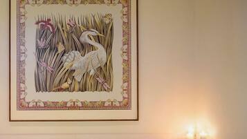 Een lijstwerk met een afbeelding van een vogel tussen rietstengels aan een muur, een bad met een paarse handdoek en twee brandende kaarsen op een plank.