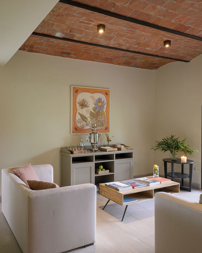 A cozy living room with a brick ceiling, a large floral painting, a grey sofa, a coffee table with books, and a side table with a plant and candle.