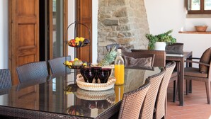 Un coin repas avec une table à plateau en verre, des chaises en osier, un panier de fruits et une bouteille de jus d’orange.