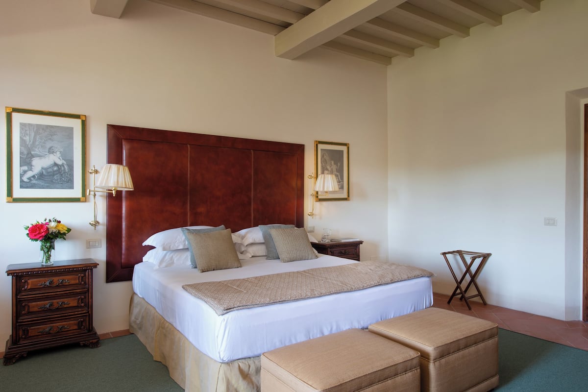 Een slaapkamer met een groot bed, een houten hoofdbord, nachtkastjes, een nachtkastje, een klein houten krukje en twee ingelijste foto’s aan de muur.