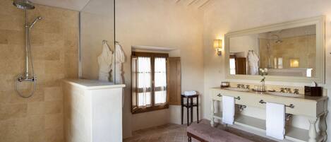 A bathroom with a shower, a bench, a vanity with a mirror, and a window with curtains.