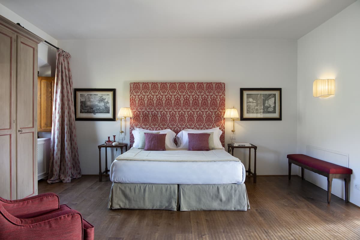 Een netjes opgemaakt slaapkamer met een groot bed, twee nachtkastjes met lampen, een bankje en twee ingelijste foto's aan de muur.