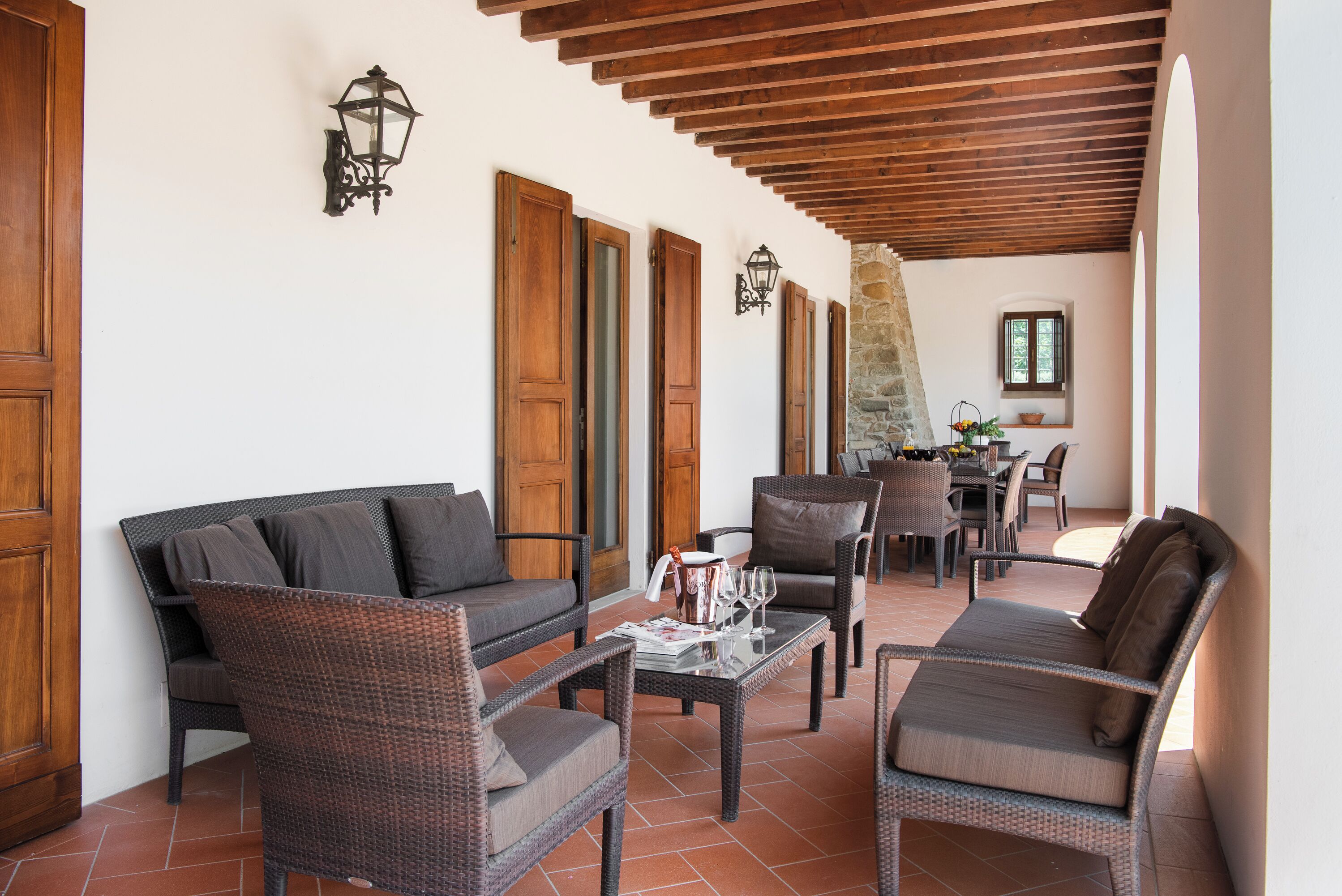 Un patio techado con muebles de mimbre, un muro de piedra y una zona de comedor con sillas y una mesa.