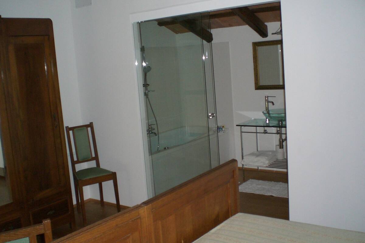 A bedroom with a wooden bed, a glass shower enclosure, a wooden chair, and a bathroom with a sink and mirror.