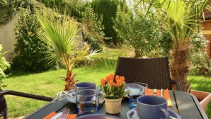 Et udendørs spisebord dækket med tallerkener, bestik og glas, omgivet af frodig vegetation og en palme.