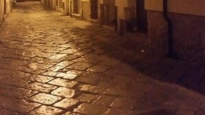 Uma estreita rua de paralelepípedos com edifícios em ambos os lados, com portas em arco e varandas com grades de metal.