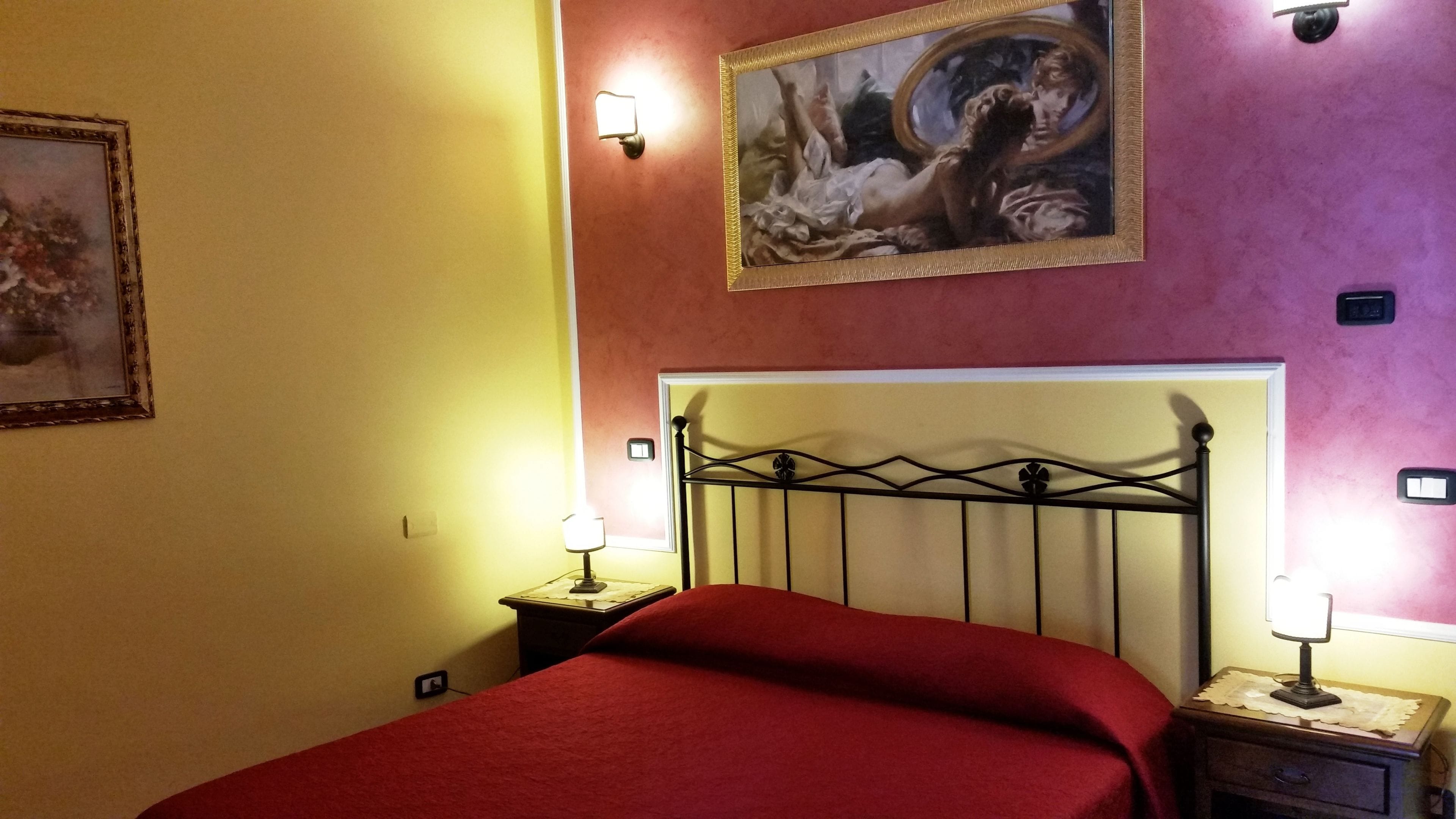 A bedroom with a bed, two bedside tables with lamps, a wall-mounted painting, and a light switch.