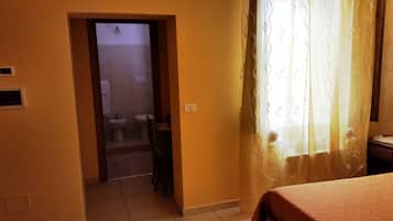 Ein Hotelzimmer mit Bett, einem durchgegangenen Tür erkennbaren Badezimmer und einem Vorhang, der ein Fenster teilweise verdeckt.