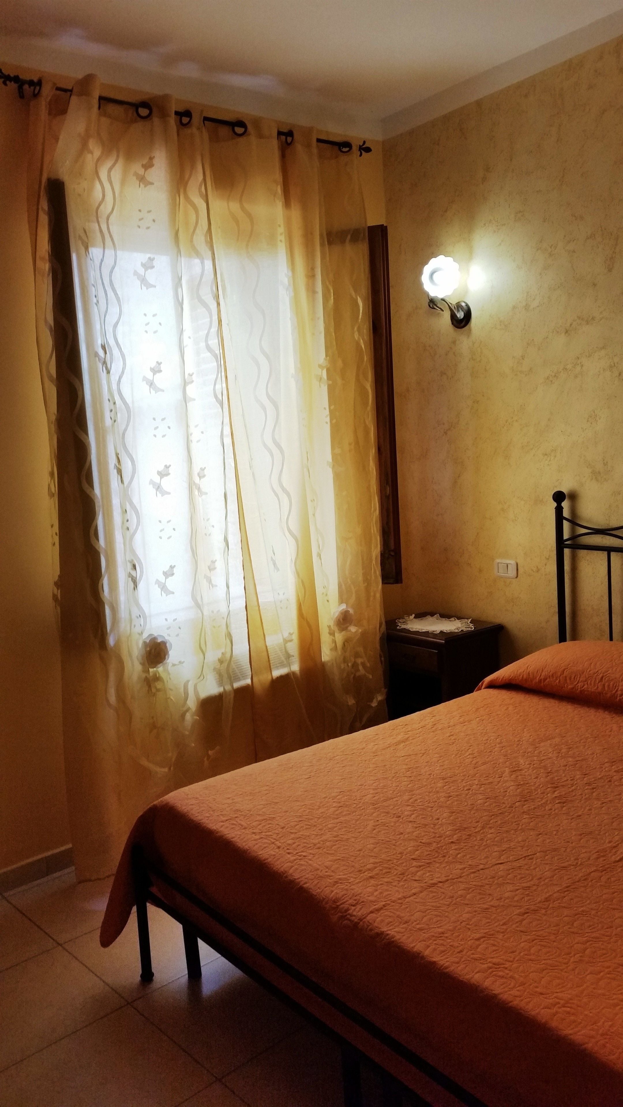 A room with a bed, a nightstand, a wall-mounted light, and a window with curtains.