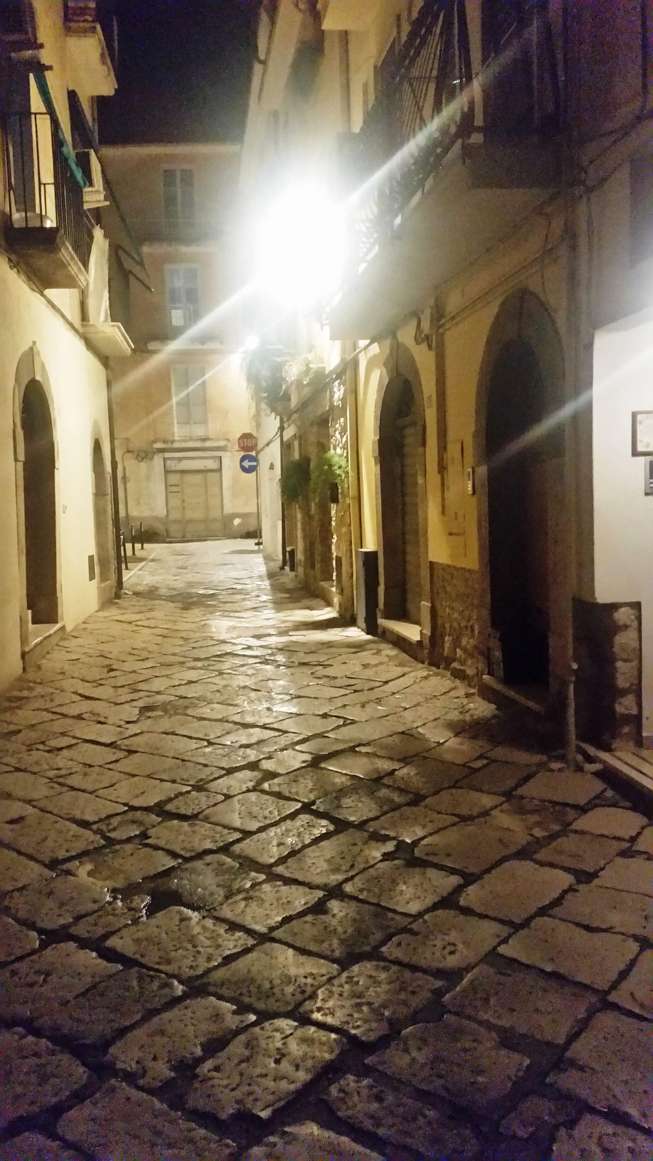 A cobblestone street at night with buildings on either side, illuminated by street lamps.