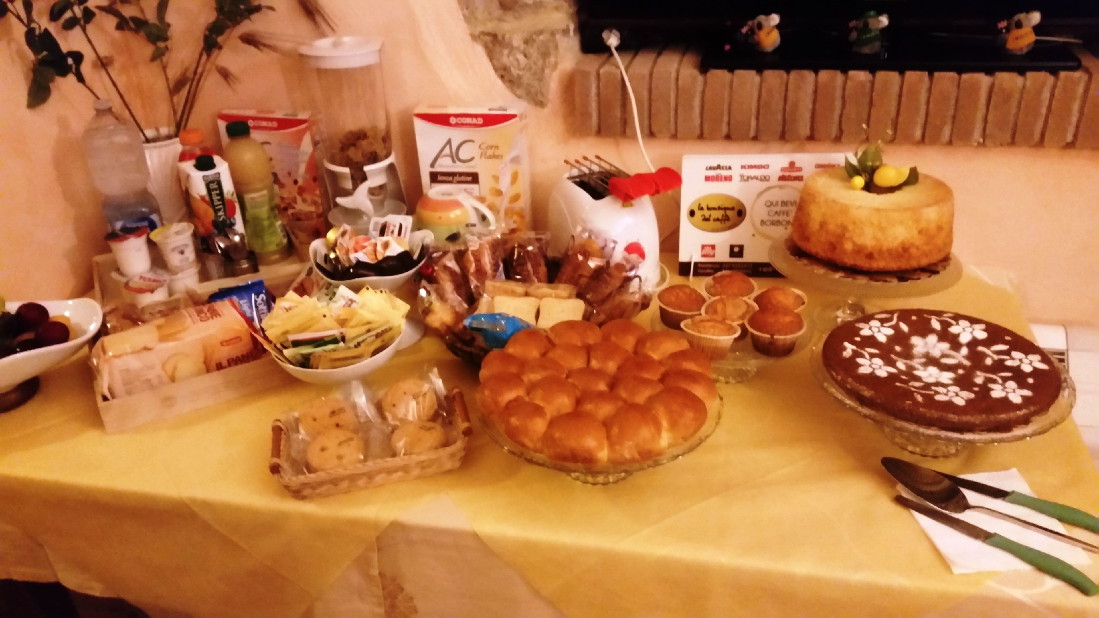 A table with various food items and a cake, including a basket of bread rolls, a bowl of fruit, and a chocolate cake with white flowers on top.
