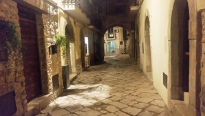 Um beco estreito de paralelepípedos com edifícios de pedra em ambos os lados, uma porta visível à esquerda e uma placa com a inscrição