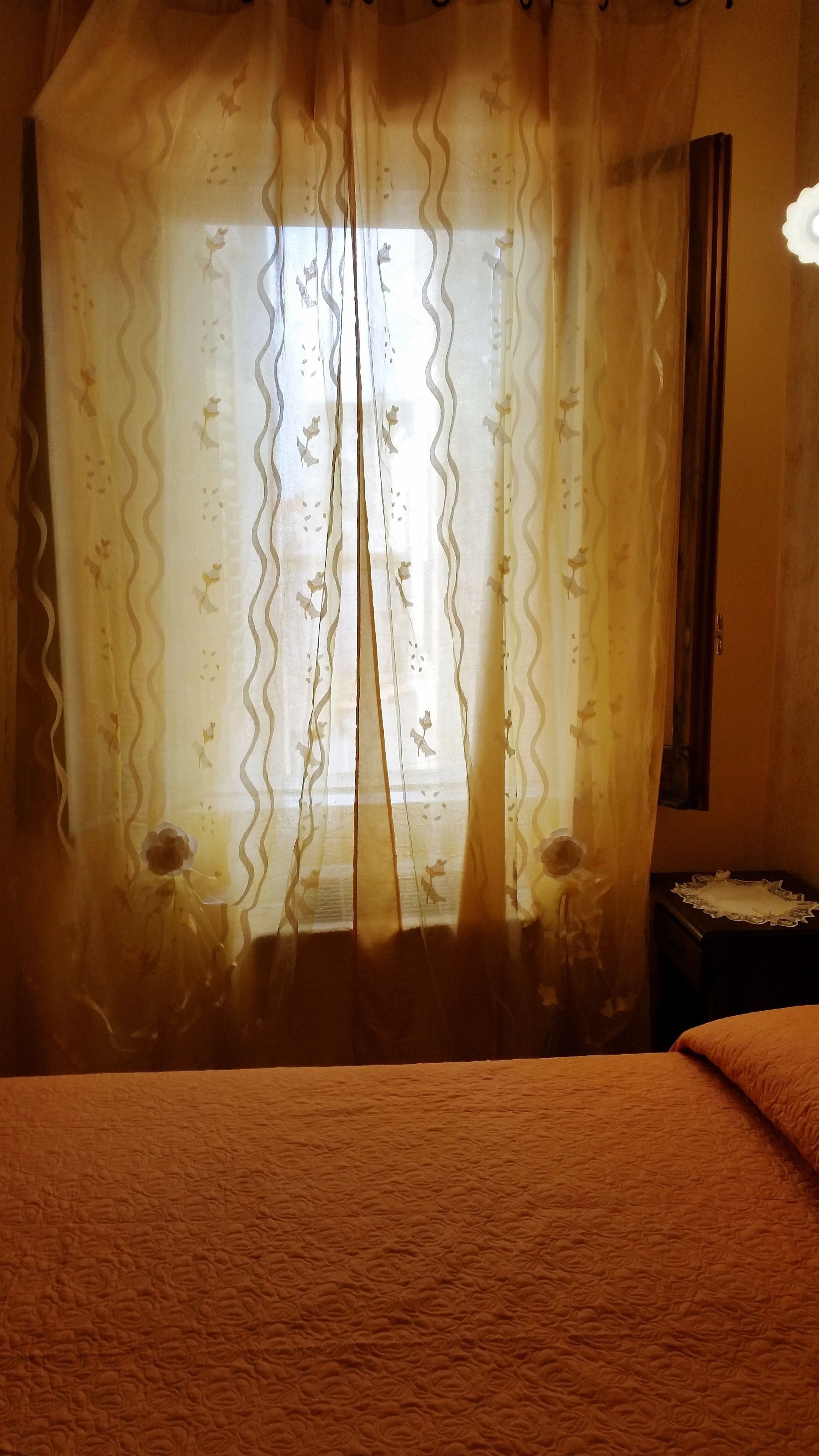 Ein Zimmer mit einem Bett, einem Fenster mit durchsichtigen Vorhängen und einem Nachttisch mit Lampe.