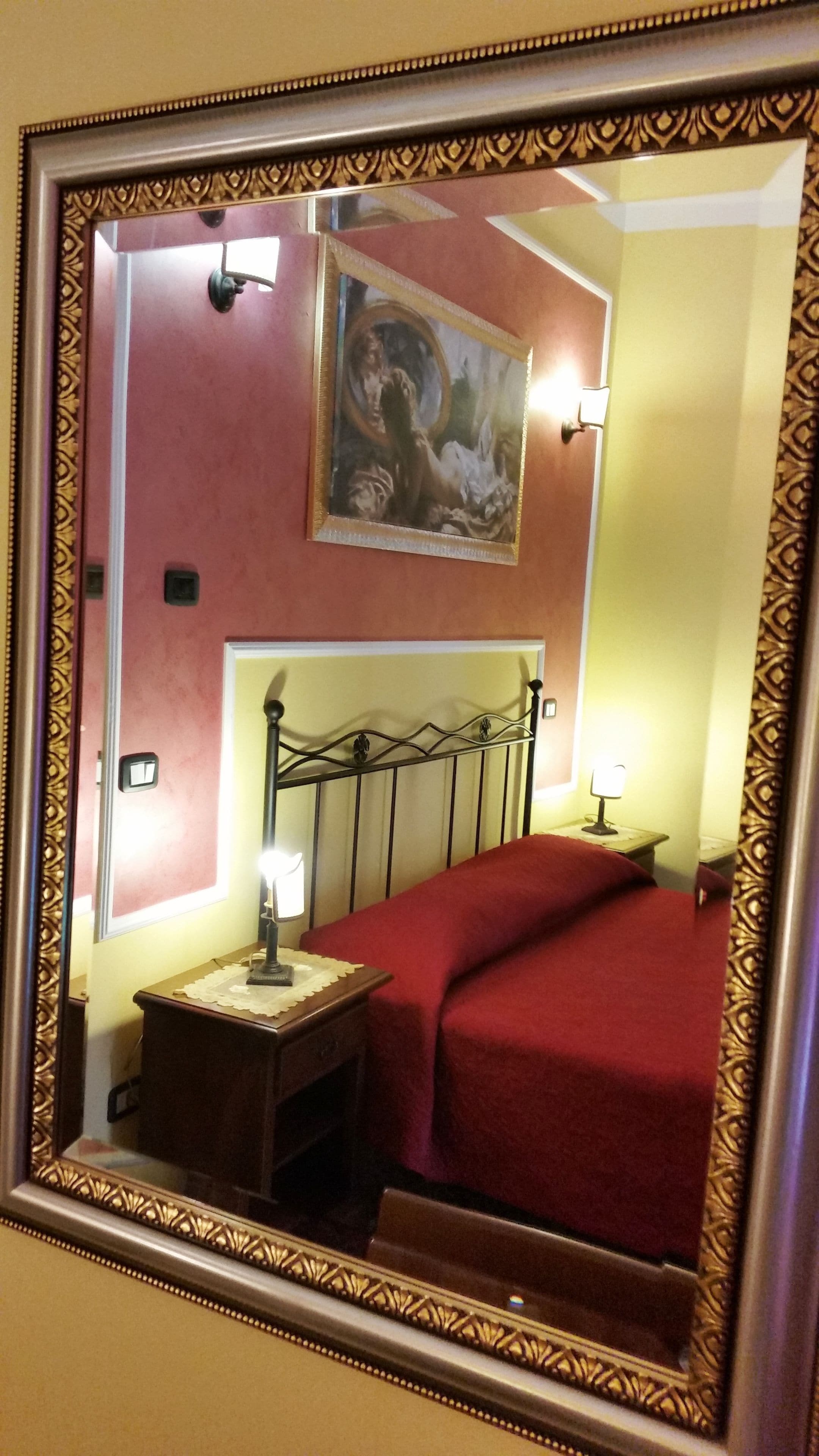 A bedroom with a bed, a nightstand, a lamp, a painting on the wall, and a mirror with an ornate frame.