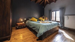 A bedroom with a wooden ceiling, a bed with a blue bedspread, yellow pillows, a nightstand with a lamp, and a window with curtains. - Agriturismo Borgoluce (Susegana)