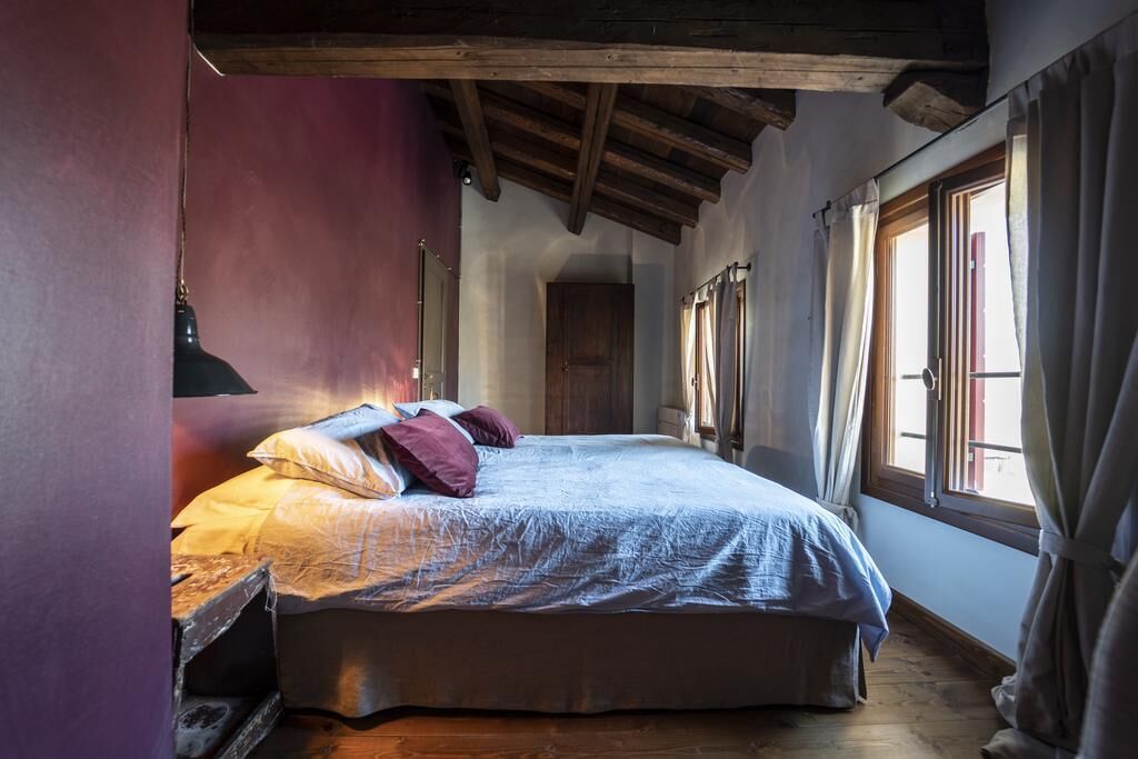 A bedroom with a large bed, wooden beams, a window with curtains, and a wooden floor.