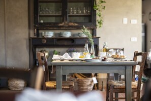 Una mesa de comedor preparada con alimentos para el desayuno, rodeada de sillas de madera y una cesta de mimbre.