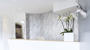 A modern reception area with a marble wall, a white reception desk, and a mounted animal head sculpture. - Alpin Life Hotel Gebhard (Fiss)
