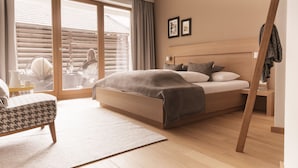 A modern bedroom with a large bed, a sitting area with a patterned chair, and a view through a large window. - Alpin Life Hotel Gebhard (Fiss)
