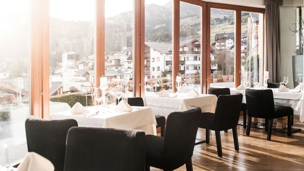 A dining area with tables set for four, large windows offering a view of a mountainous village. - Alpin Life Hotel Gebhard (Fiss)