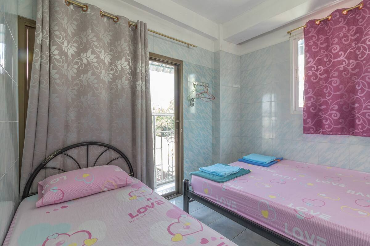 A bedroom with two beds, a window with curtains, and a bathroom with a shower curtain.