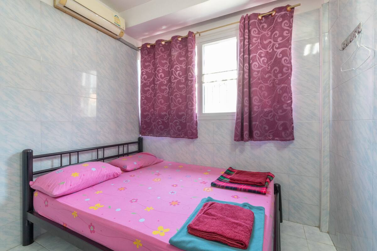 A single bed with pink bedding, a window with purple curtains, and a wall-mounted air conditioner.