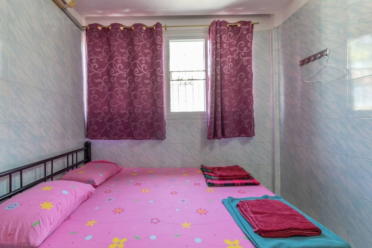 A single bed with a floral bedspread, pink pillows, and a red towel on a blue bed skirt.